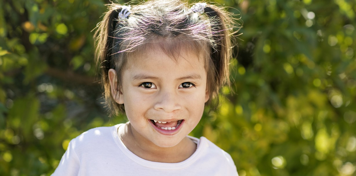 Three-year-old Karla shares her beautiful new smiles 15 months after her cleft lip surgery. Karla is outside wearing pigtails in her hair and a white shirt as she stands in front of wall of greenery. Photo: Jasmin Shah.