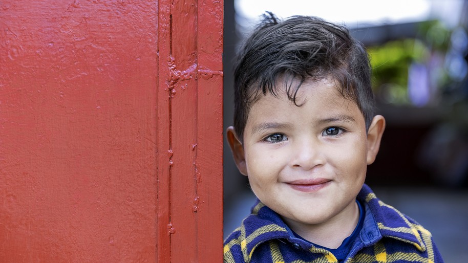 Wearing a yellow and blue long-sleeved flannel shirt, Dilan stands outside his red front door of his home and shows off his new smile two years after receiving cleft lip surgery. 