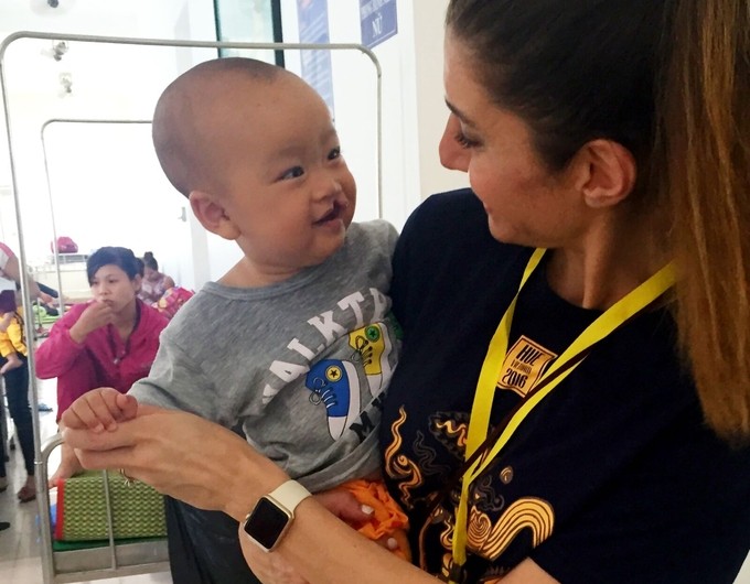 Dr. Neda Kalantar, pediatric dentist, shares a smile small child with a cleft lip during a past Operation Smile surgical program. Neda is wearing a black T-shirt with a yellow lanyard, and the child is wearing a grey T-shirt as Neda holds them, making eye contact and smiling. 