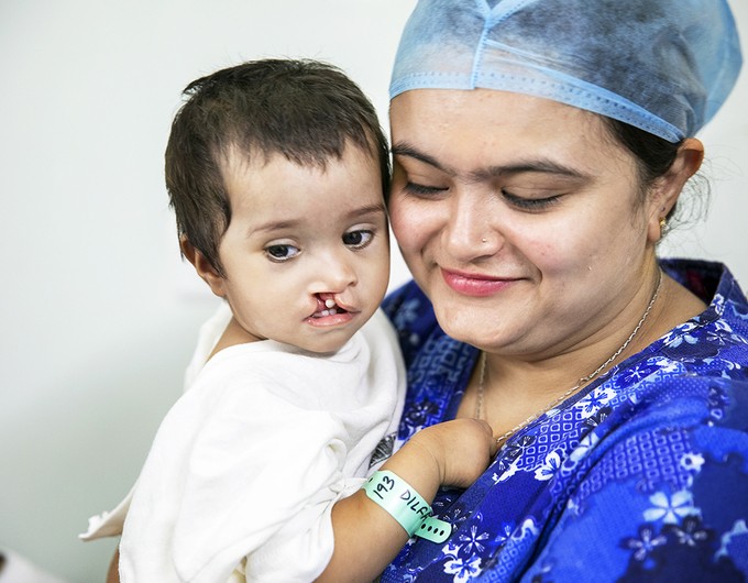 Six-month-old Dilfa wears a white surgical gown while being comforted by a medical volunteer in blue scrubs during an Operation Smile India surgical program.