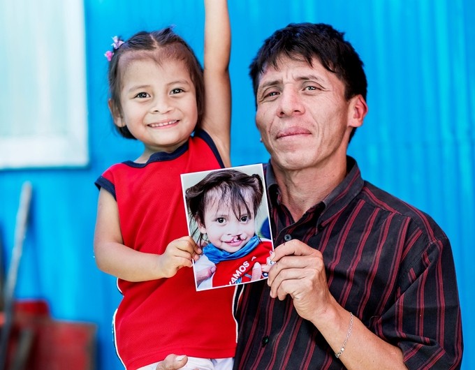 Karla, in a red shirt, sits in the arm of her father, Dede, with her arm shooting up toward the skies while Dede holds her and a photo of her before surgery. Photo: Jasmin Shah.