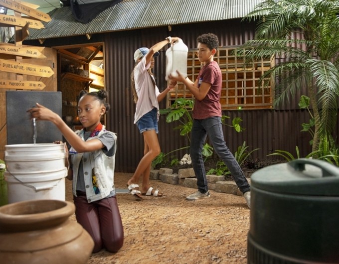 Children engage with the Interactive Learning Center. The exhibit replicates a rural village in a low- or middle-income country. A girl observes a fixture that shows water flowing into a bucket, which two children lift a water jub in the background.