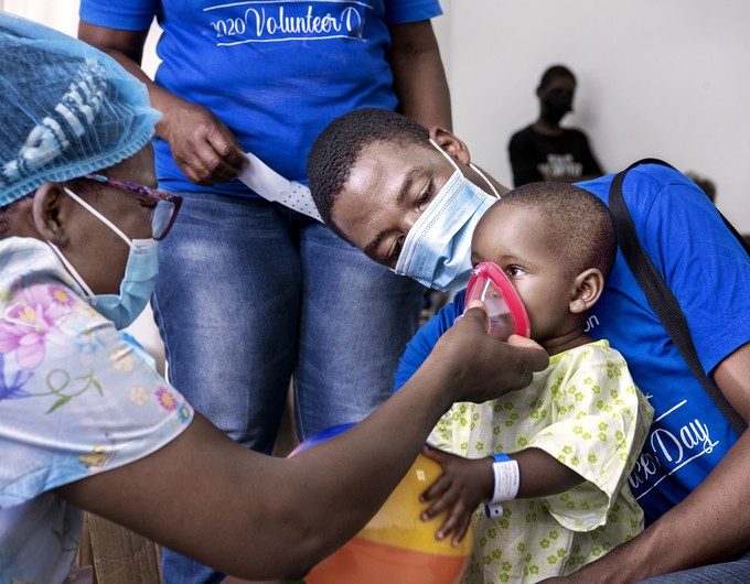 Wearing a surgical gown, Julita plays with a ball in the arms of pre- and post-operative nurse Donald Mlombwa of Malawi while psycosocial care provider Cathy Cheonga of Malawi helps them feel familiar with an anesthesia mask during an Operation Smile Malawi surgical program in 2022. 