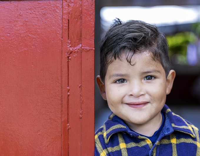 Wearing a yellow and blue long-sleeved flannel shirt, Dilan stands outside his red front door of his home and shows off his new smile two years after receiving cleft lip surgery. 