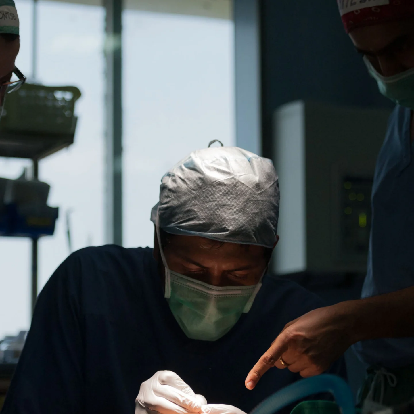 Dr. Nicolas Sierra performs surgery under the direction of Plastic Surgeon Hiteshwar Sarma. They wear protective masks and caps perform a procedure over an unshown patient in a hospital setting, with one clinician leaning in while another reaches toward an instrument or equipment.