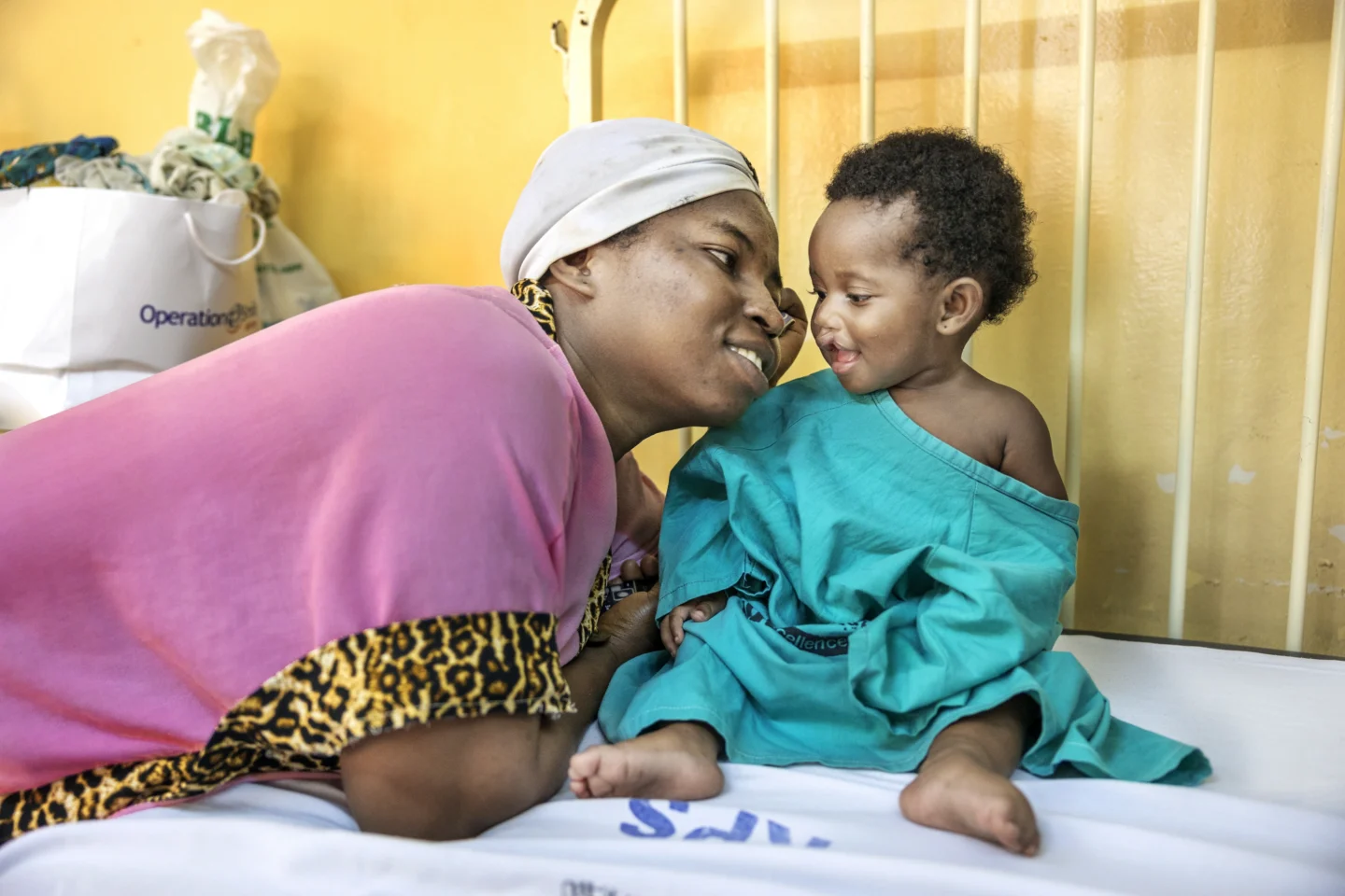 Farida sits upright on a bed, smiling at her mother, Alimatu, who is smiling and leaning close to Farida.