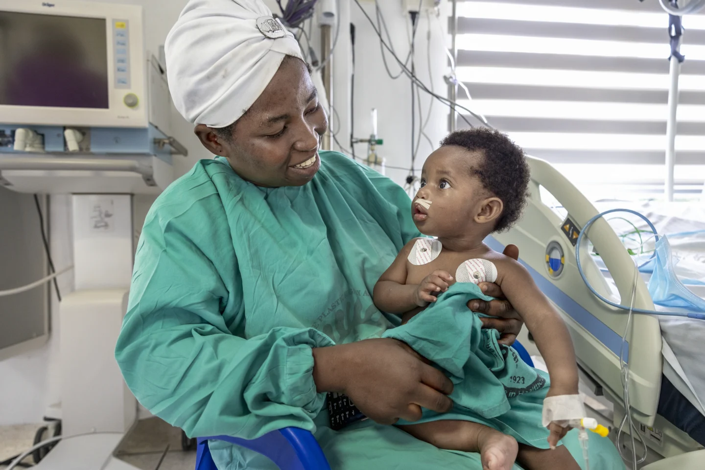 Alimatu wears a surgical gown while holding her daughter Farida post-surgery. Farida is awake and looking at her mother. They are sitting in a hospital bed and Farida's vitals are being monitored by nearby equipment.