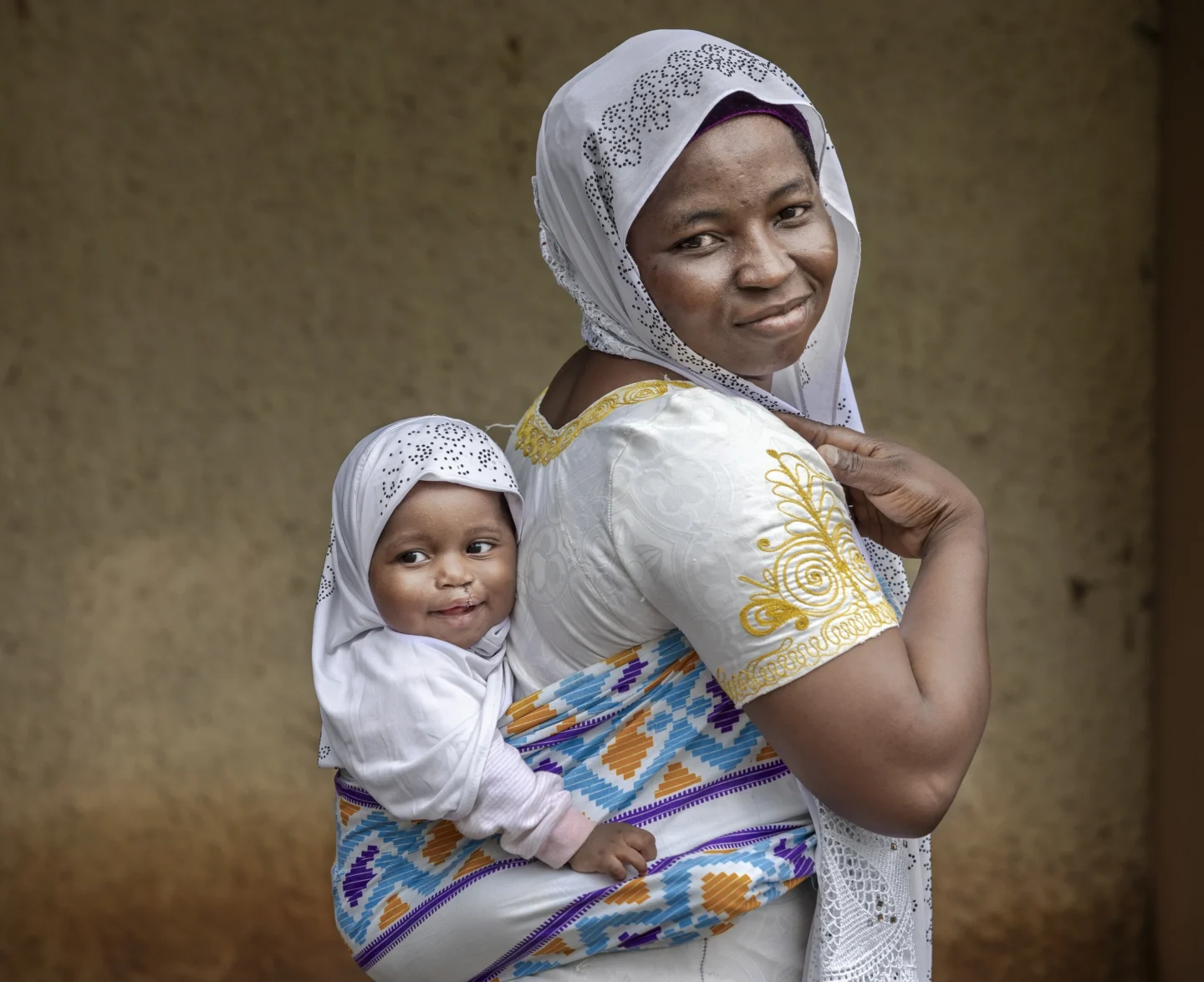 Alimatu smiles and looks over her shoulder at the camera, Farida, recovered from her surgery, is carried behind her with a wrap. Farida is smiling and looking off camera.
