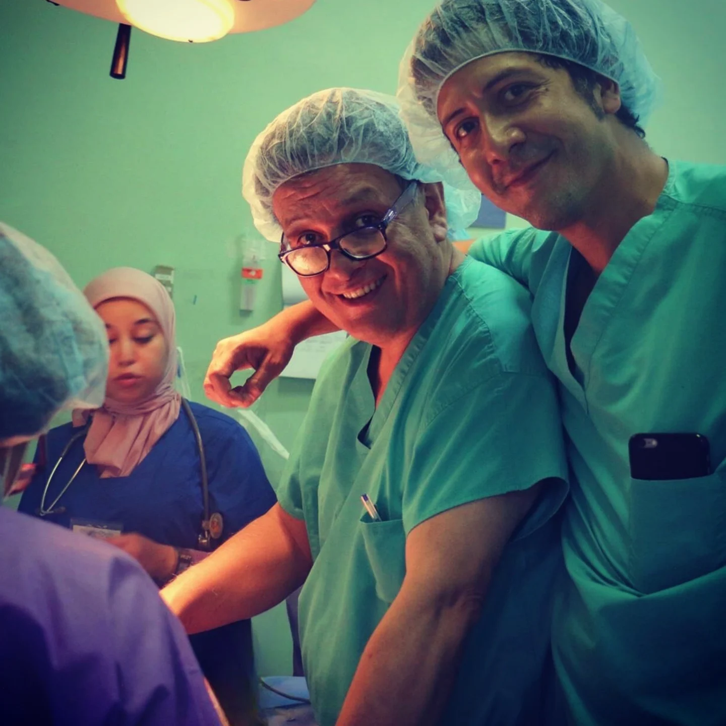 Dr. Nicolas Sierra takes a selfie-style photo with his father, Dr. Plinio Sierra. They are smiling at the camera while wearing green scrubs and disposable hair covers in an operating room.