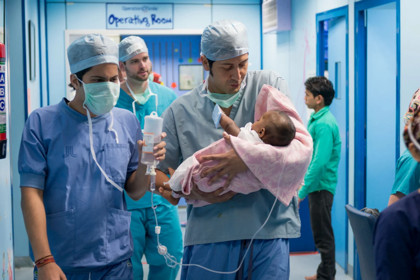 Dr. Nicolas Sierra carries a newborn wrapped in a pink blanket through a hospital corridor, with medical staff and equipment visible in the background. He wears blue scrubs and is accompanied by other surgeons in blue scrubs and surgical masks.