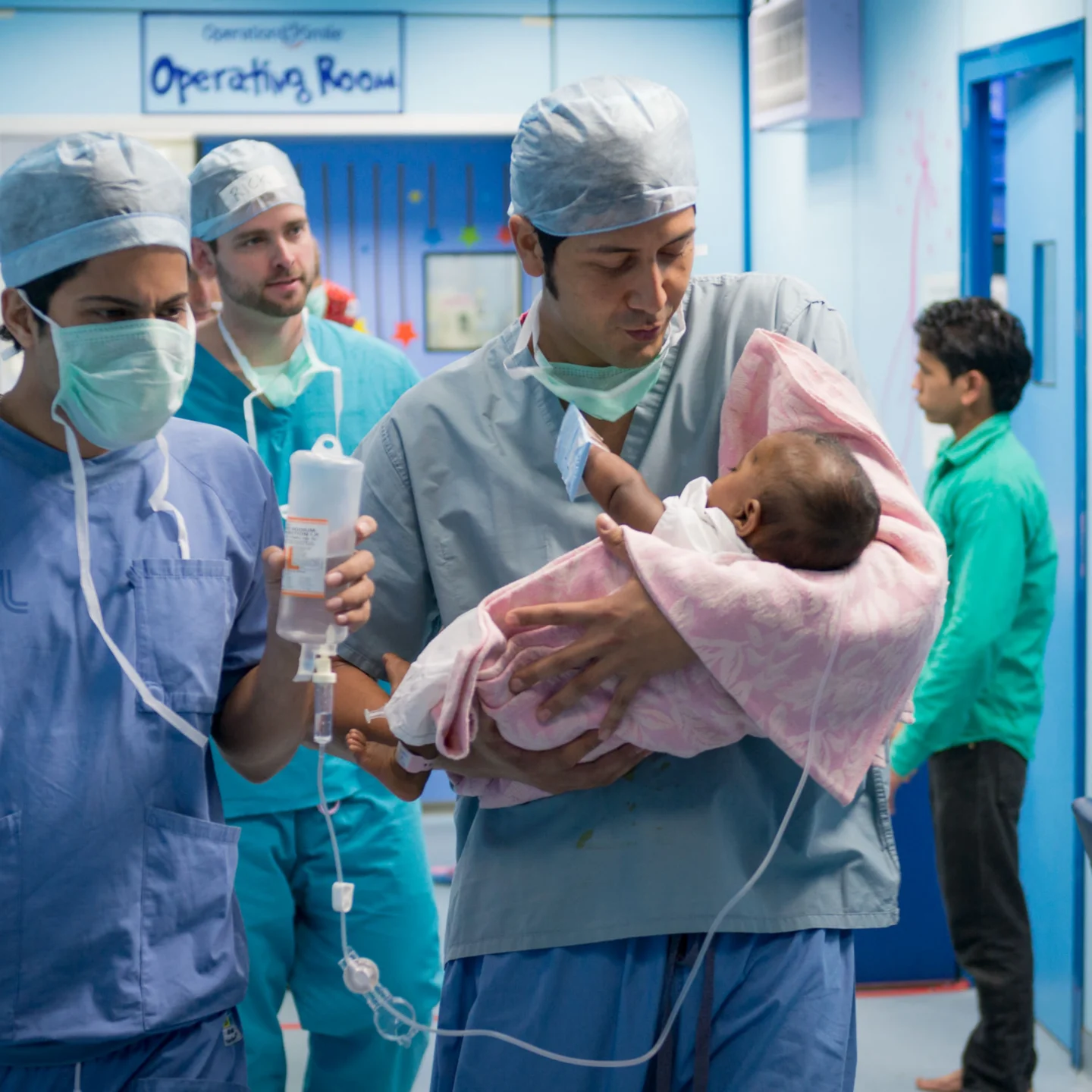 Dr. Nicolas Sierra carries a newborn wrapped in a pink blanket through a hospital corridor, with medical staff and equipment visible in the background. He wears blue scrubs and is accompanied by other surgeons in blue scrubs and surgical masks.