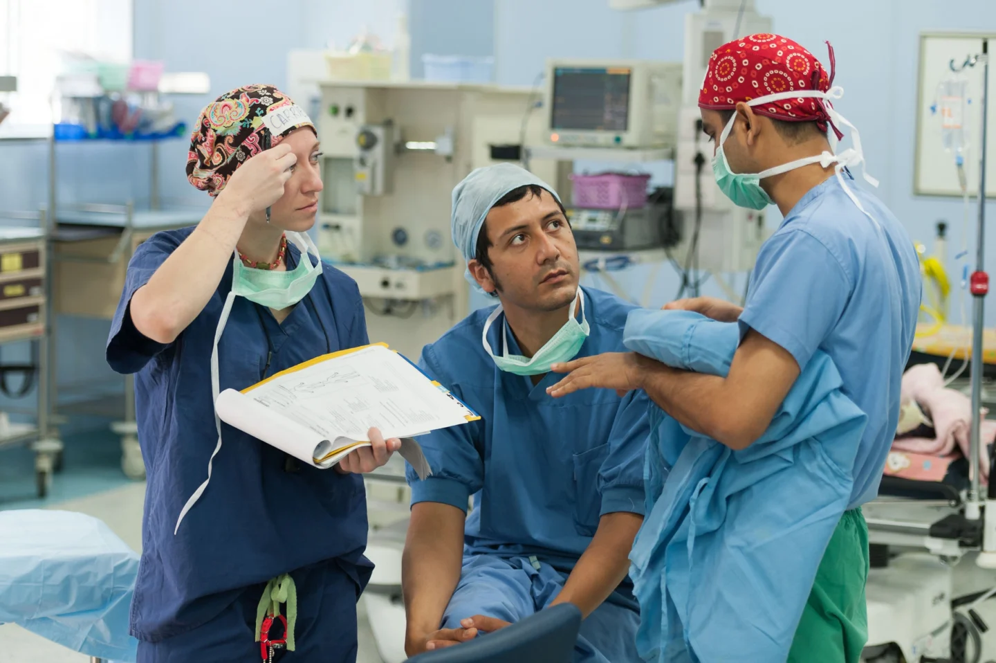 Dr. Nicolas Sierra confers with other medical staff. They wear blue scrubs and surgical caps and stand in a hospital operating room, one holding an open clipboard.