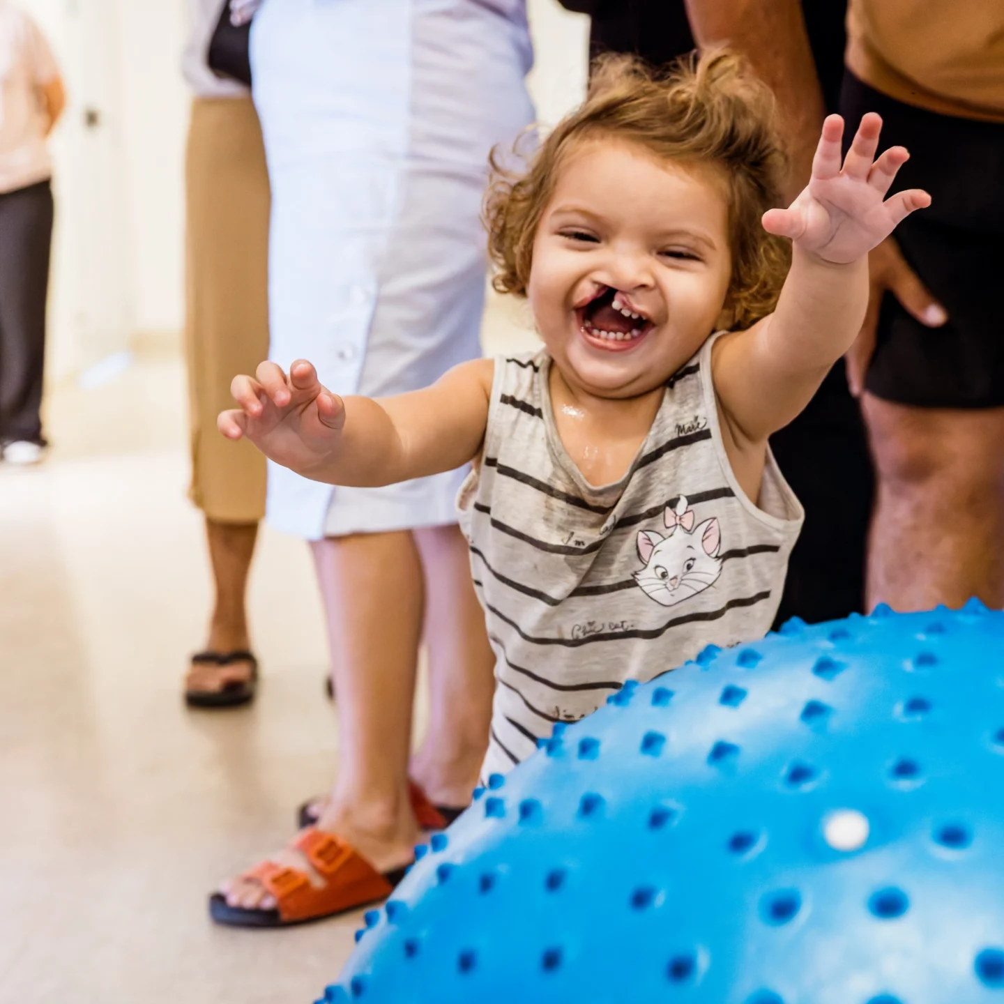 A smiling Isis wears a striped sleeveless shirt and raises both hands while standing indoors near a large blue balance ball with dotted texture.