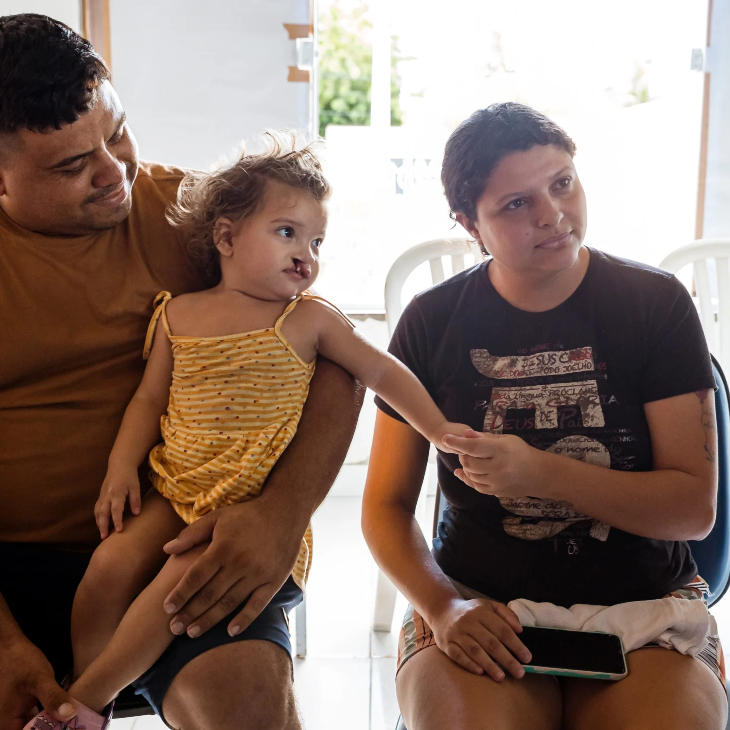 Sitting in her father's lap, Isis reaches for her mother Erica, who sits beside her listening to a healthcare provider. They are seated indoors with bright light from a window behind them.