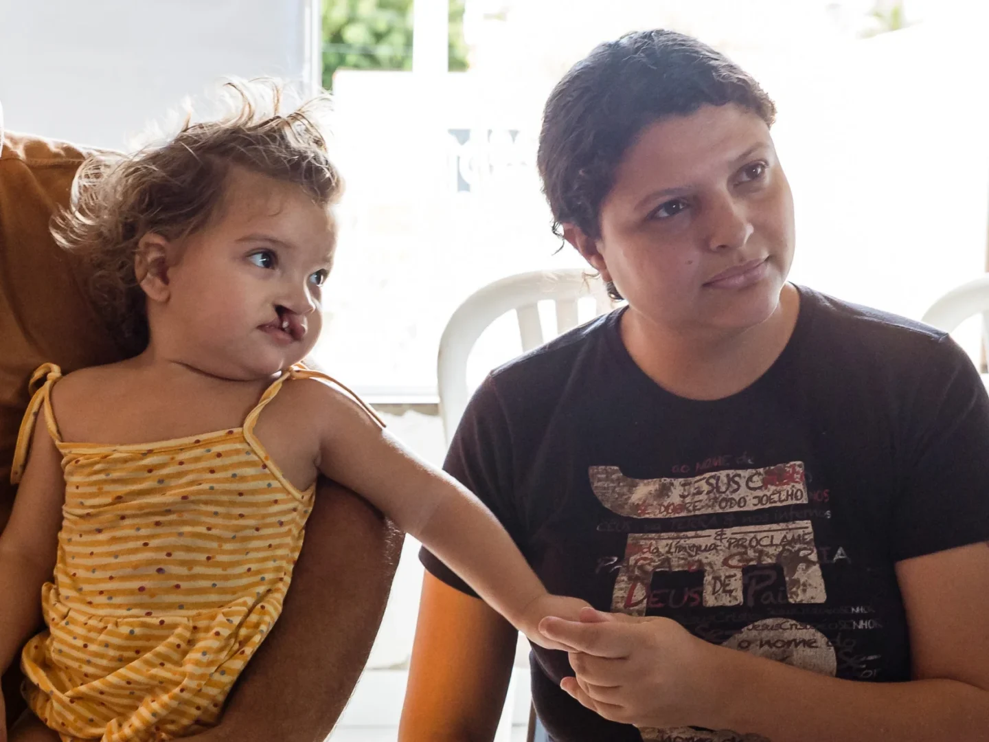 Sitting in her father's lap, Isis reaches for her mother Erica, who sits beside her listening to a healthcare provider. They are seated indoors with bright light from a window behind them.