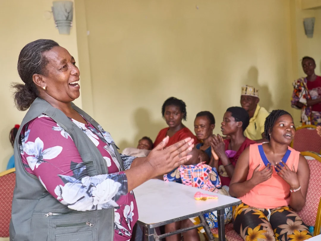 Nancy stands at the front of a classroom smiling and clasping her hands in front of a group of men and women in Kenya who are seated watching her presentation. Some of the listeners are holding infants in their arms or laps. The listeners are talking to each other as well, suggesting an energetic discussion among the group.