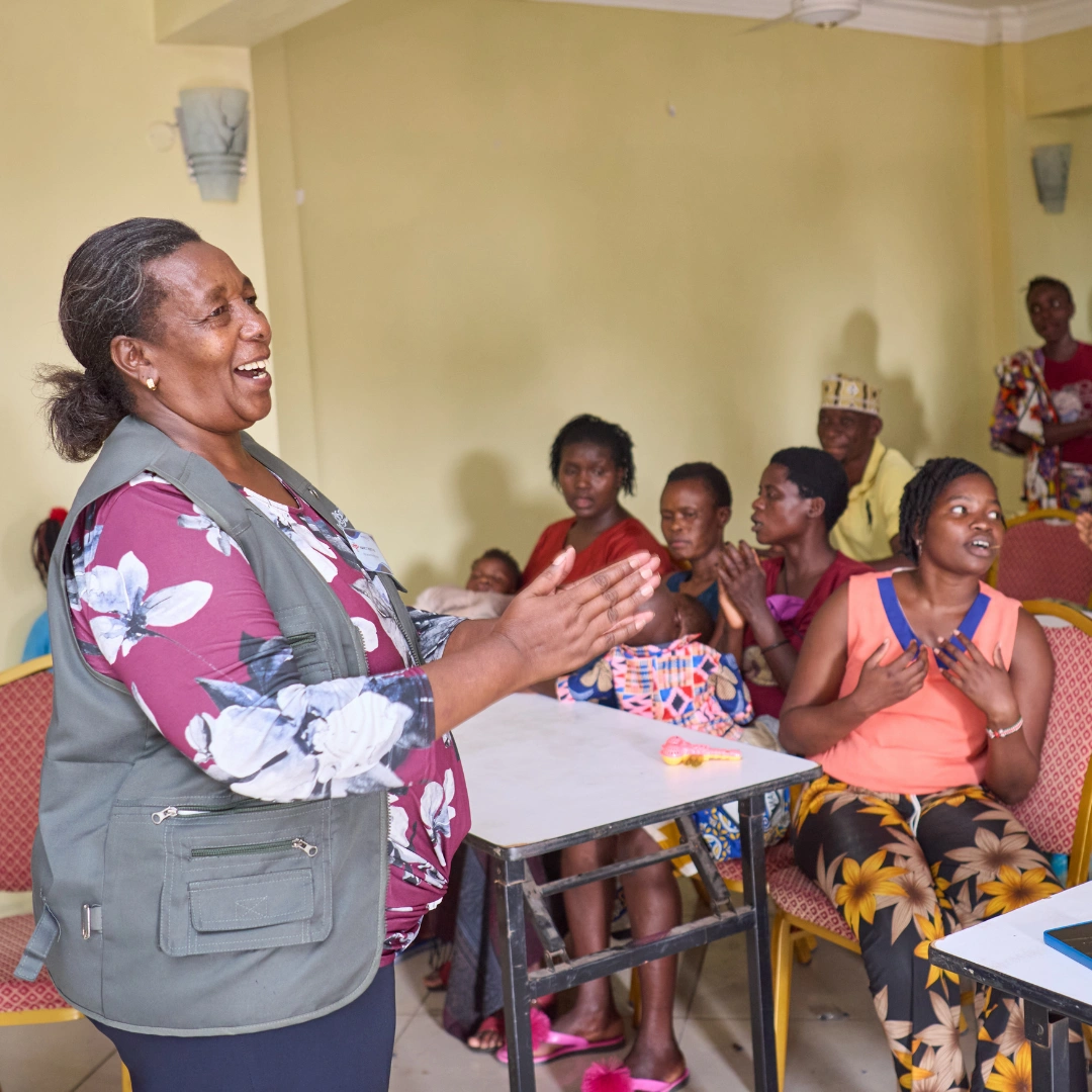 Nancy stands at the front of a classroom smiling and clasping her hands in front of a group of men and women in Kenya who are seated watching her presentation. Some of the listeners are holding infants in their arms or laps. The listeners are talking to each other as well, suggesting an energetic discussion among the group.