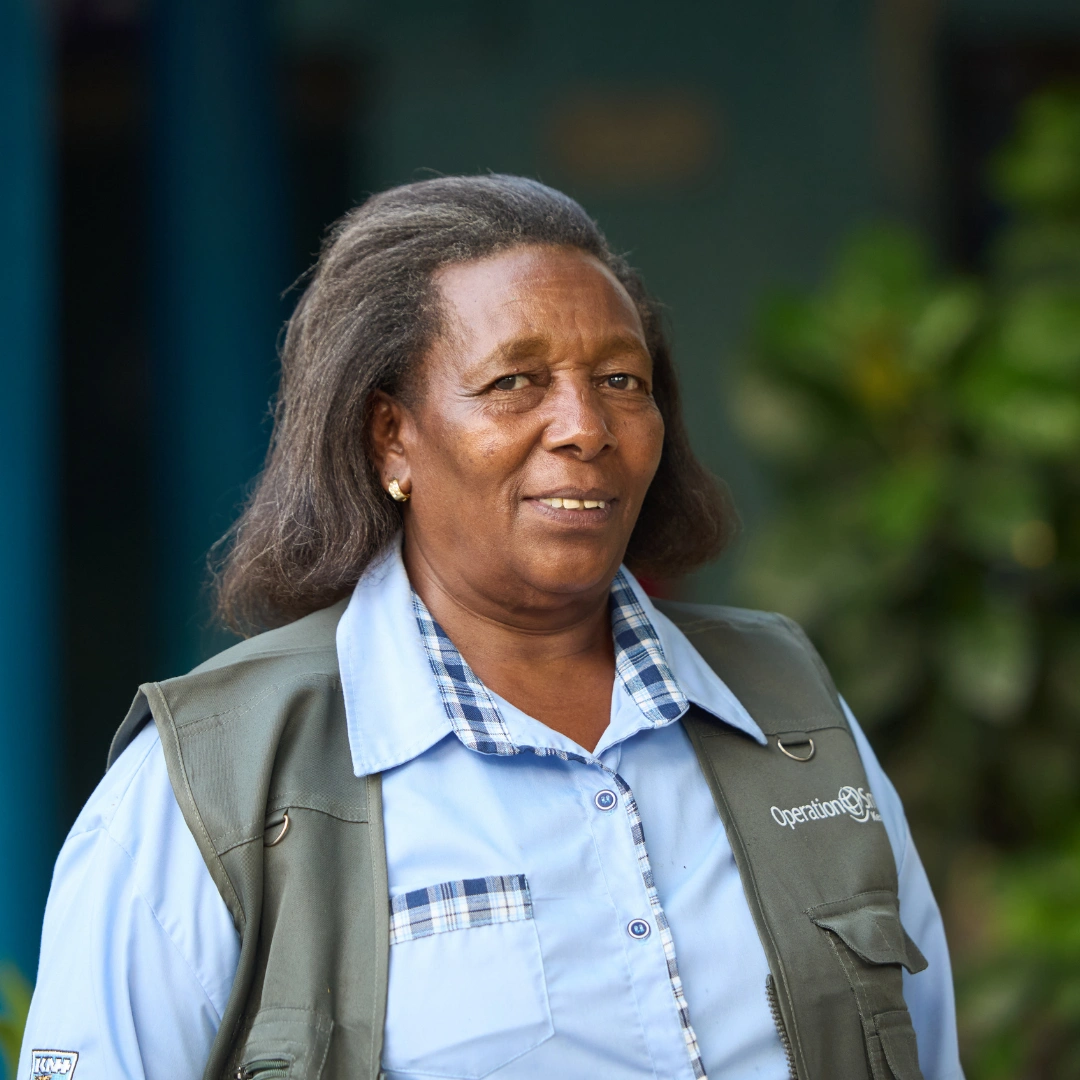 A picture of Nancy, a psychosocial care worker in Kenya, smiling at the camera.. Nancy is a middle-aged woman with shoulder-length dark hair with gray streaks, and she is wearing a light blue button-up shirt with blue-checkered design elements and a muted green vest that has the Operation Smile logo on it in white.