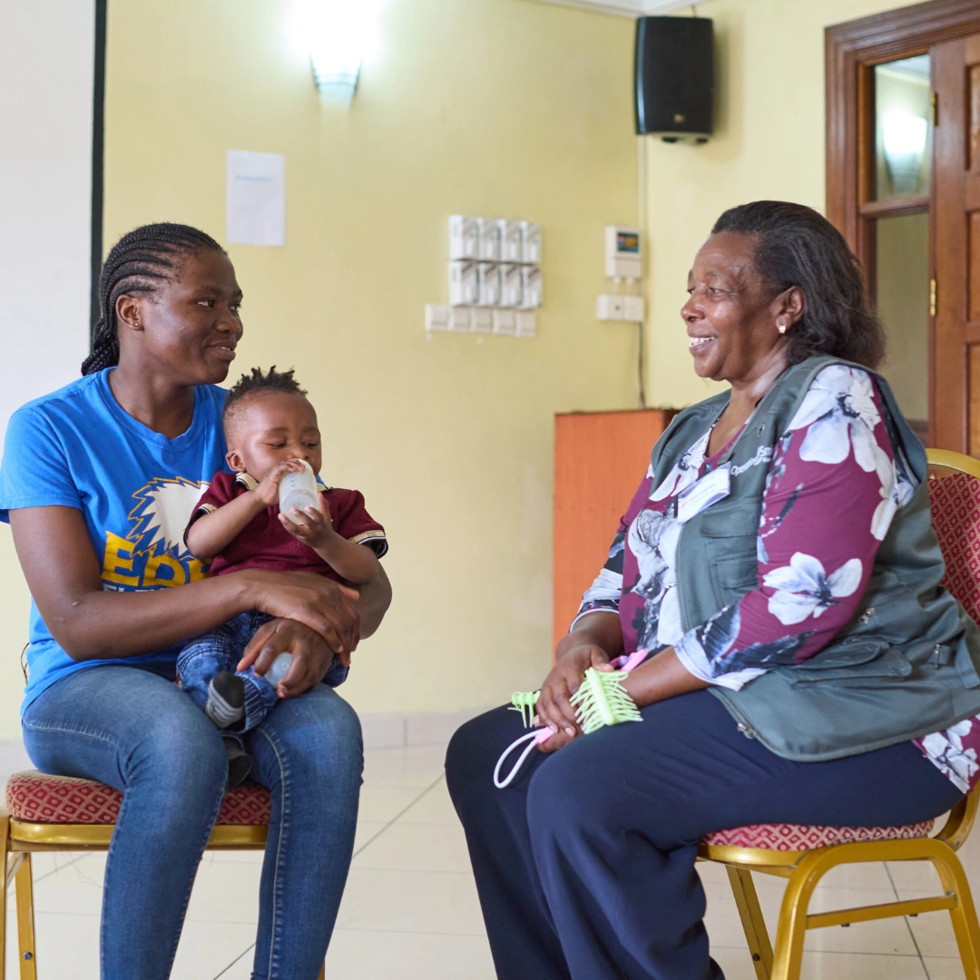 Nancy, a middle-aged woman from Kenya, sits across from a mother holding her infant, who is drinking from a bottle. They are smiling and talking in an empty classroom with a large white board, yellow wall, and glass door behind them.