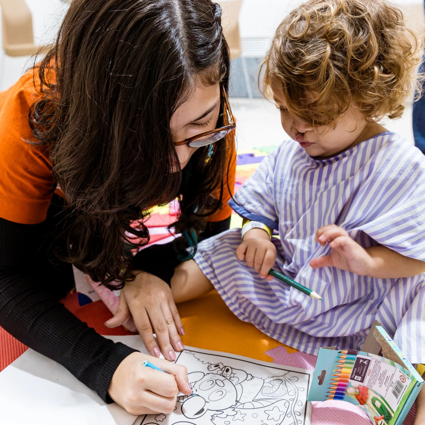 A young woman with long brown hair and glasses sits beside Isis at a table coloring a large coloring book, with crayons in hand and a colorful classroom setting in the background.
