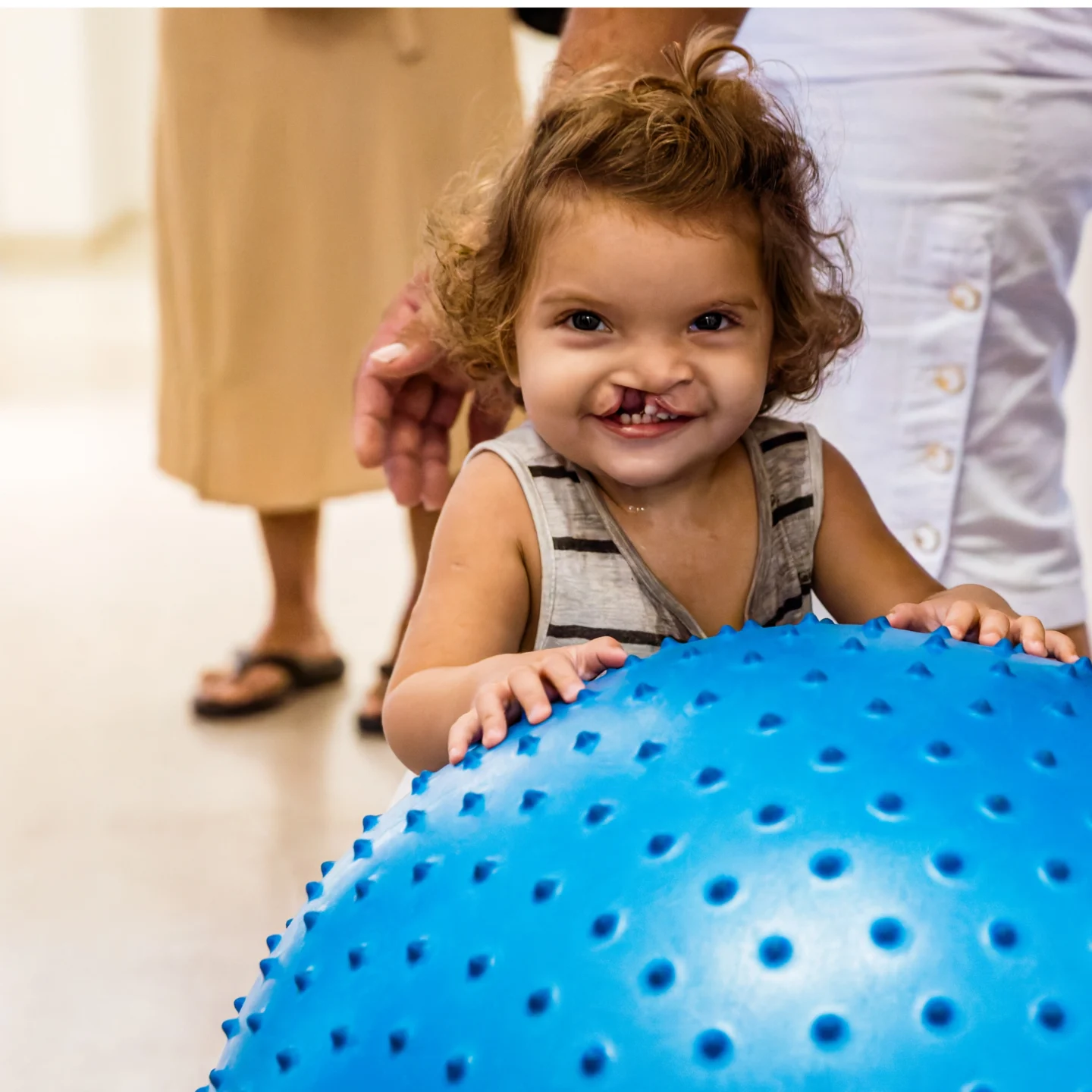 Isis smiles and holds onto a large blue exercise ball with people around her in a bright room.