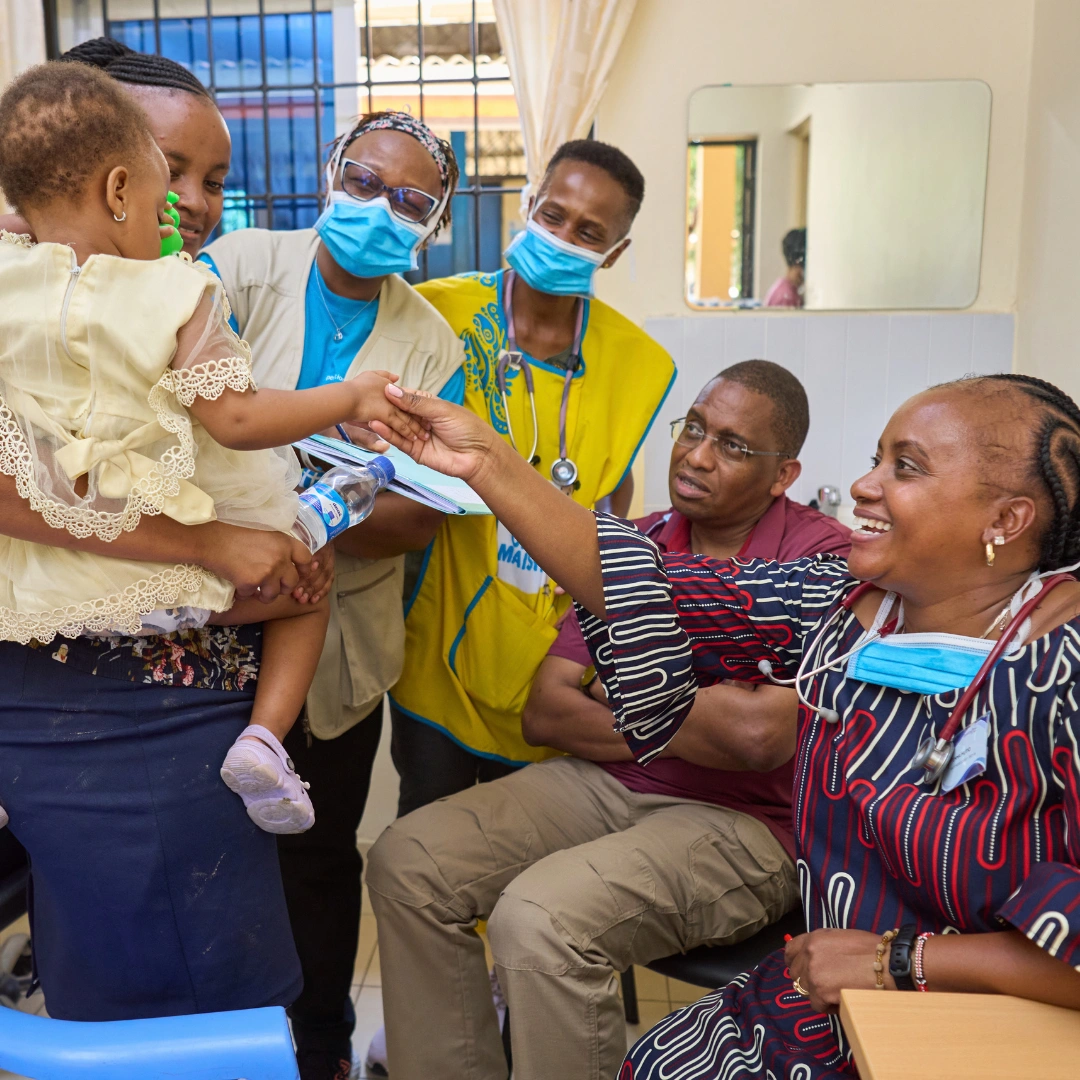 A child in a clinic in Kenya reaches toward a nurse wearing a blue mask, while a doctor and seated patient watch; colorful medical walls and chairs are in the background.