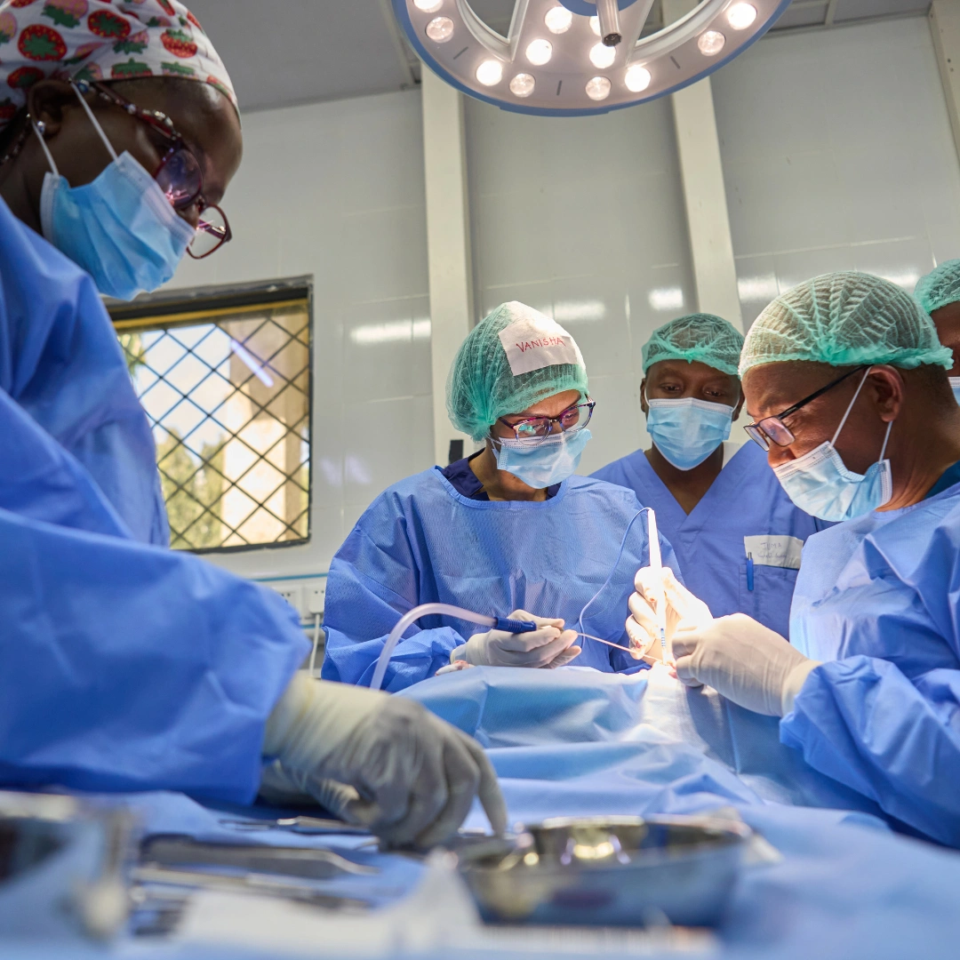 A group of surgeons in blue surgical gowns, masks, and caps perform an operation in a bright operating room, leaning over an open surgical field while overhead lights illuminate the scene.