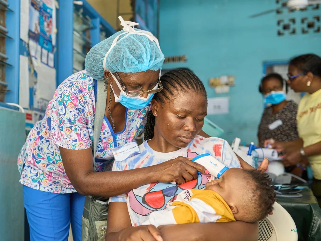 A healthcare worker in Kenya, wearing colorful scrubs and a surgical hairnet and facemask, leans over to check on an infant resting in its mother's arms while she sits on a chair.
