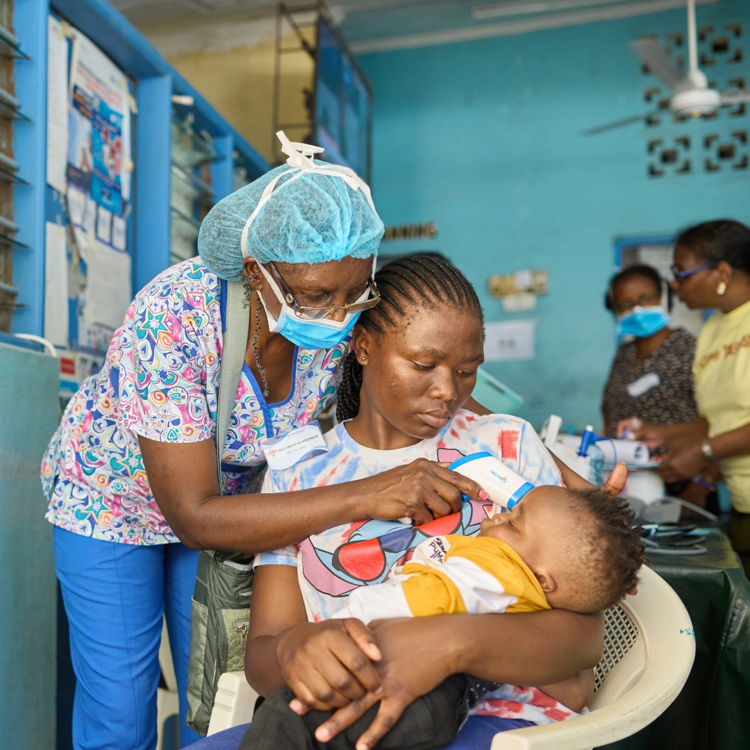 In a brightly lit clinic with turquoise walls, a healthcare worker in Kenya, wearing colorful scrubs and a surgical hairnet and facemask, leans over to check on an infant resting in its mother's arms while she sits on a chair.