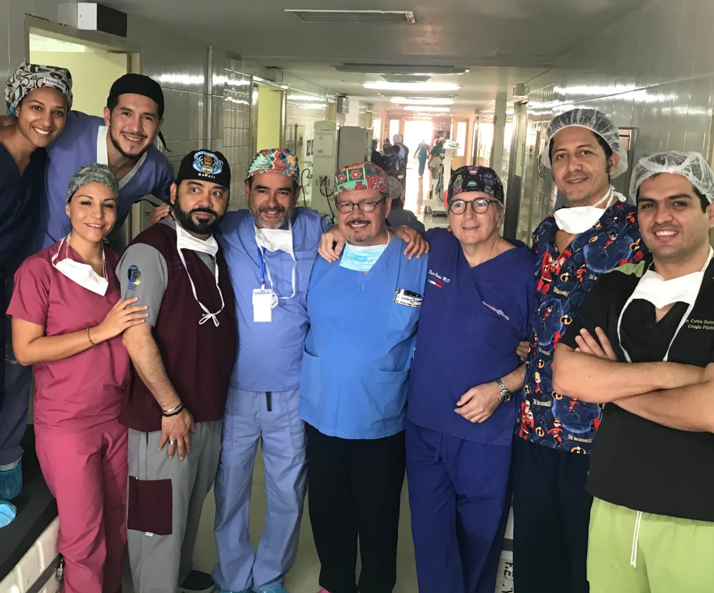 Dr. Nicolas Sierra stands with a group of healthcare workers in scrubs and head coverings posing together in a hospital hallway, smiling and standing close to the camera.
