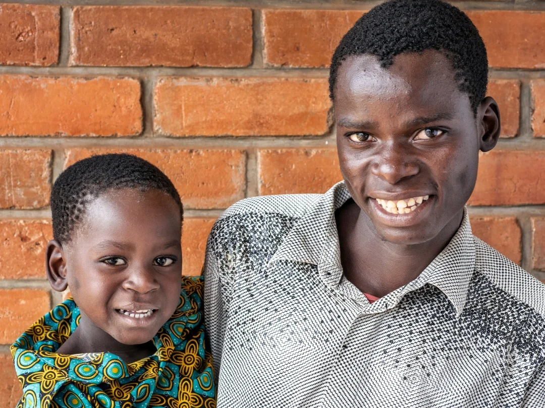 Mphatso and his father from Malawi pose together in front of a brick wall. Mphatso wears a patterned blue-and-yellow shirt, while his father wears a black-and-white patterned button-down shirt.