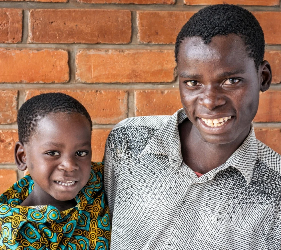 Mphatso and his father from Malawi pose together in front of a brick wall. Mphatso wears a patterned blue-and-yellow shirt, while his father wears a black-and-white patterned button-down shirt.