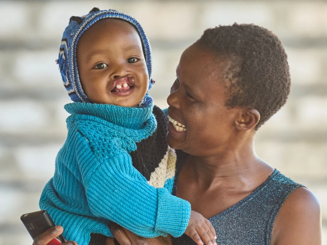 Lukas' mother smiles emotionally while holding Lukas up near her shoulder. Lukas wears a knitted bright blue sweater and cap and smiles at the camera.