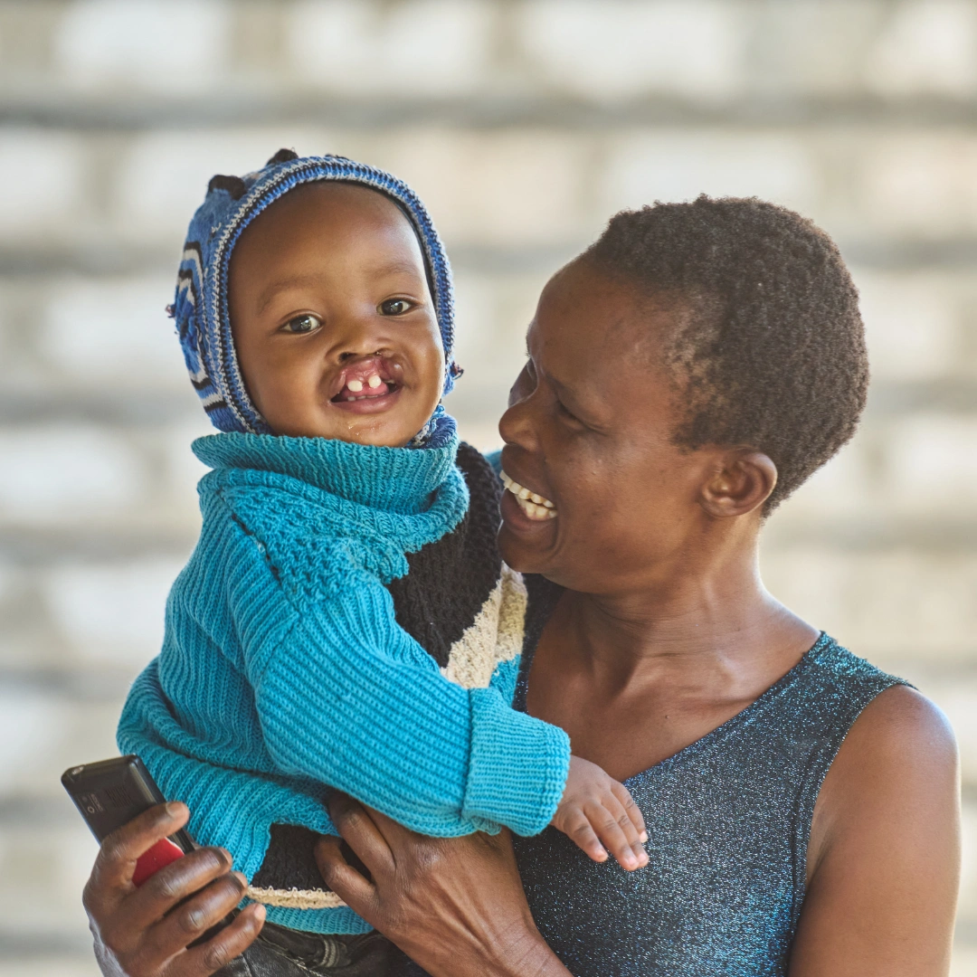Lukas' mother smiles emotionally while holding Lukas up near her shoulder. Lukas wears a knitted bright blue sweater and cap and smiles at the camera.