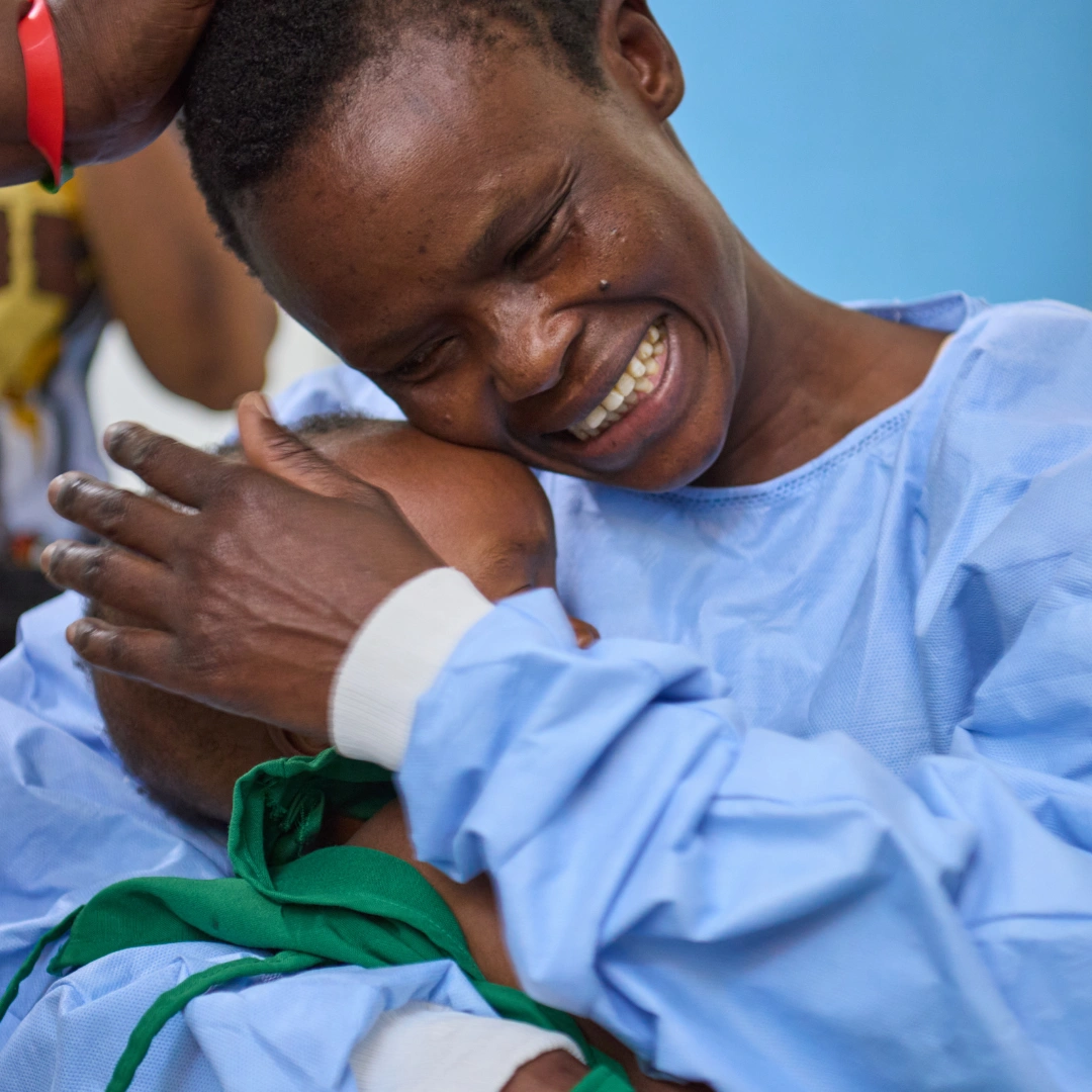 Lukas' mother wears a blue surgical gown over her clothes while she holds Lukas in a tight embrace after he has received cleft surgery.