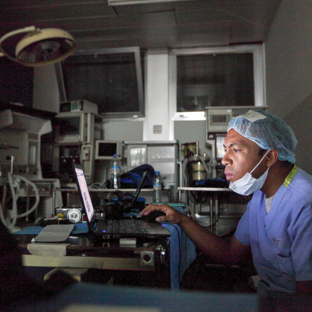 A doctor in Madagascar examines information on a laptop. The doctor wears surgical scrubs and sits in a room full of medical equipment.