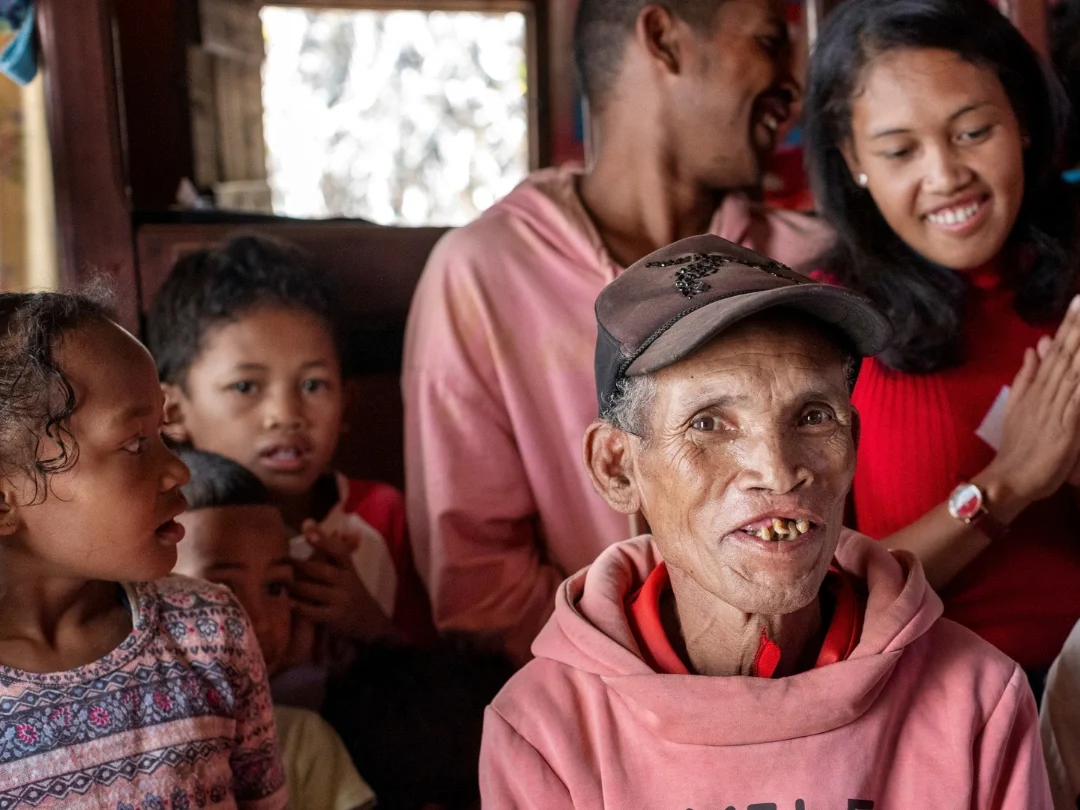 A family in Madagascar sit inside a house with brown walls. An elderly man from Madagascar smiles at the camera, to his right are three young children and behind him stand two young adults laughing and smiling.