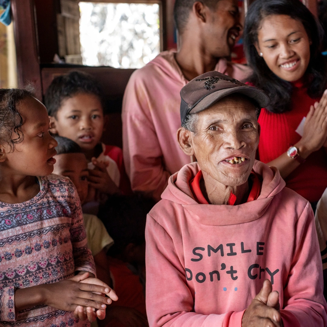 A family in Madagascar sit inside a house with brown walls. An elderly man from Madagascar smiles at the camera, to his right are three young children and behind him stand two young adults laughing and smiling.