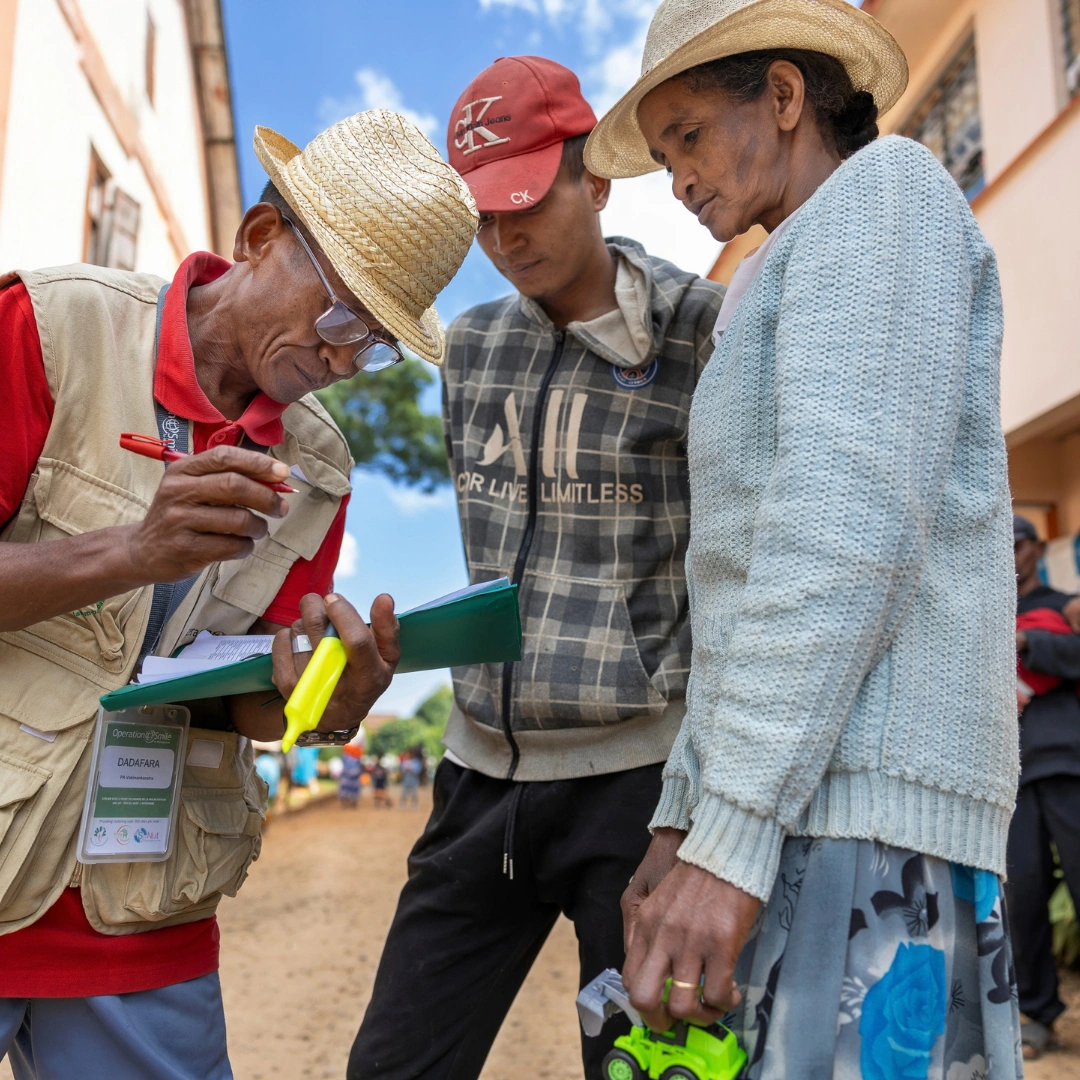An Operation Smile volunteer from Madagascar fills out a form while speaking to a man and an elderly woman. They stand outside surrounded by houses on a sunny day with blue sky. All three individuals are focused on the form being filled out.