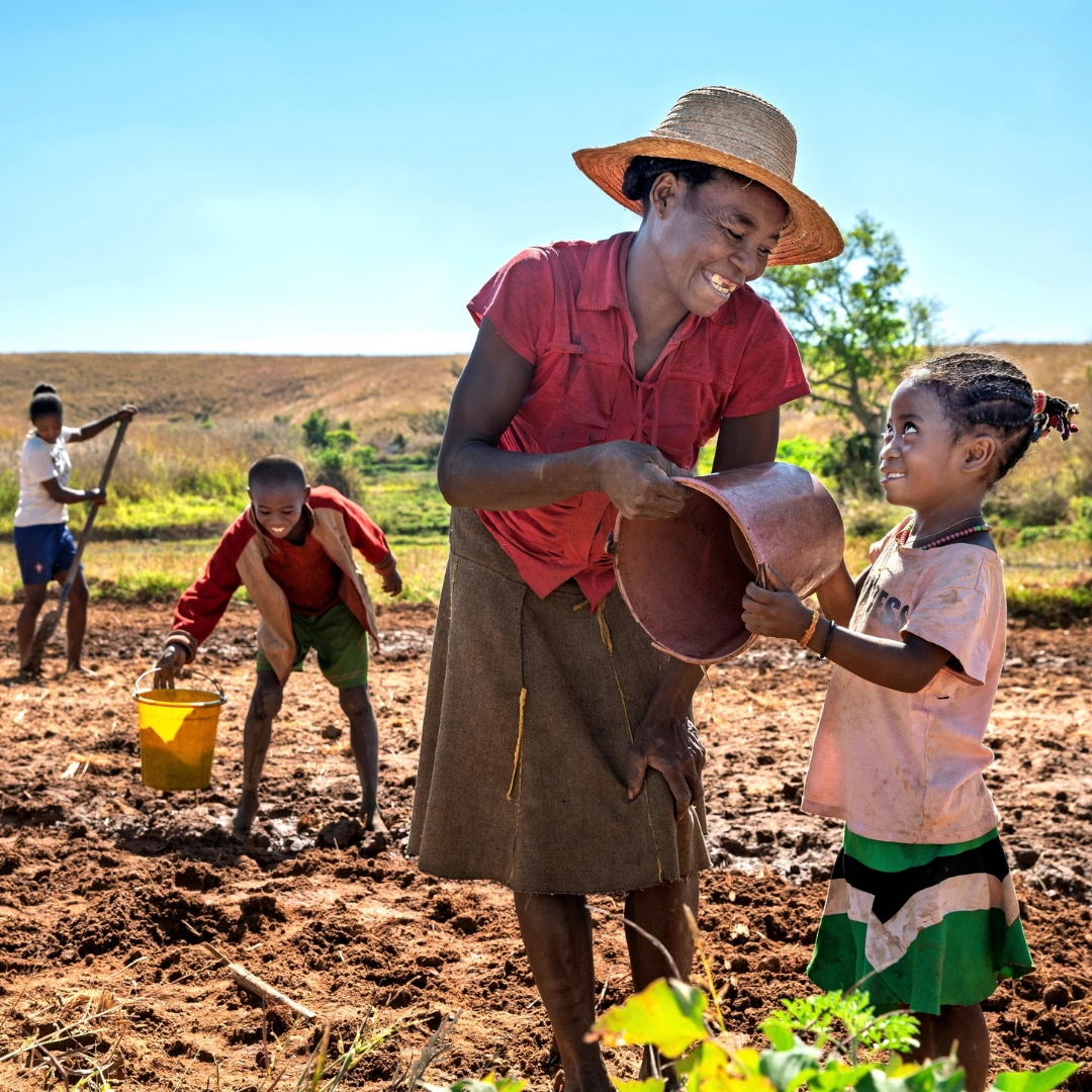 A family working on a farm in Madagascar. A woman with a straw hat and red shirt smiles at a young girl who is helping her hold a metal bucket. Behind them a boy and girl smile while working. They stand in a sunlit field with a blue sky behind them.