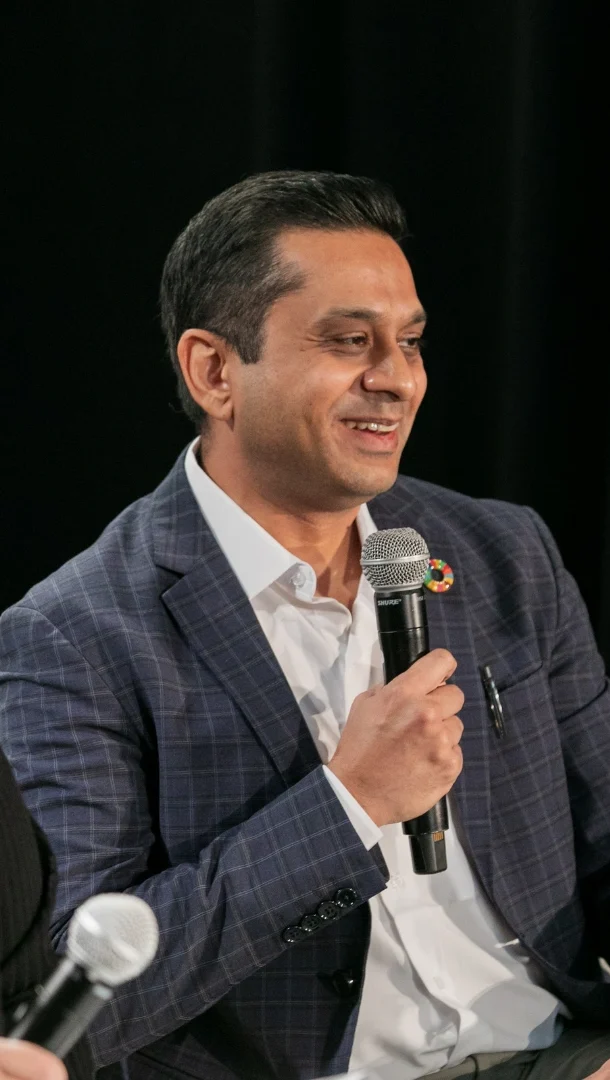 Dr. Gaurav Deshpand holds a microphone and looks off camera while speaking on a panel at an event. He wears a gray suit jacket and white collared shirt.